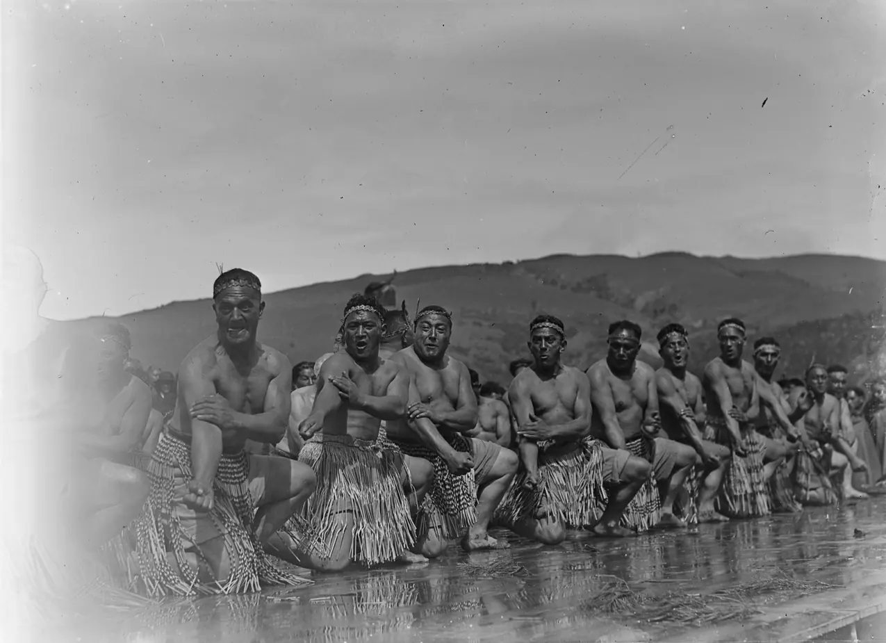 A line of men in traditional dress kneeling in front of hills, a cultural performance group photographed in the 1940s